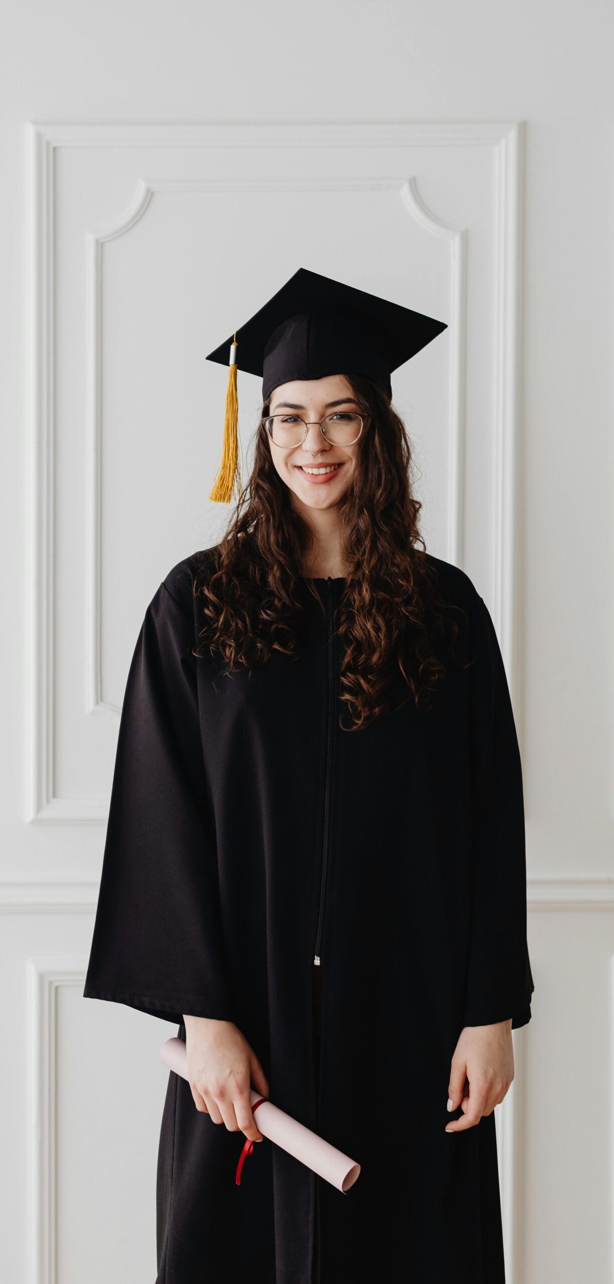 Cheerful woman in graduation attire holding diploma against white wall, symbolizing achievement.
